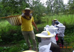 Dzieci w ubraniach pszczelarskich oraz gospodarz z plastrami miodu.